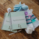 This image shows The NICU Notebook®, a coil bound NICU Journal for families with a baby in the Neonatal Intesive Care Unit, surrounded by baby items. Arranged on a wooden table top with the notebooks are a  blue crochet octopus, white nasal aspirator, purple binky, mint green pacifier, baby bottle, white micropreemie diaper, and blank neonatal hospital band.