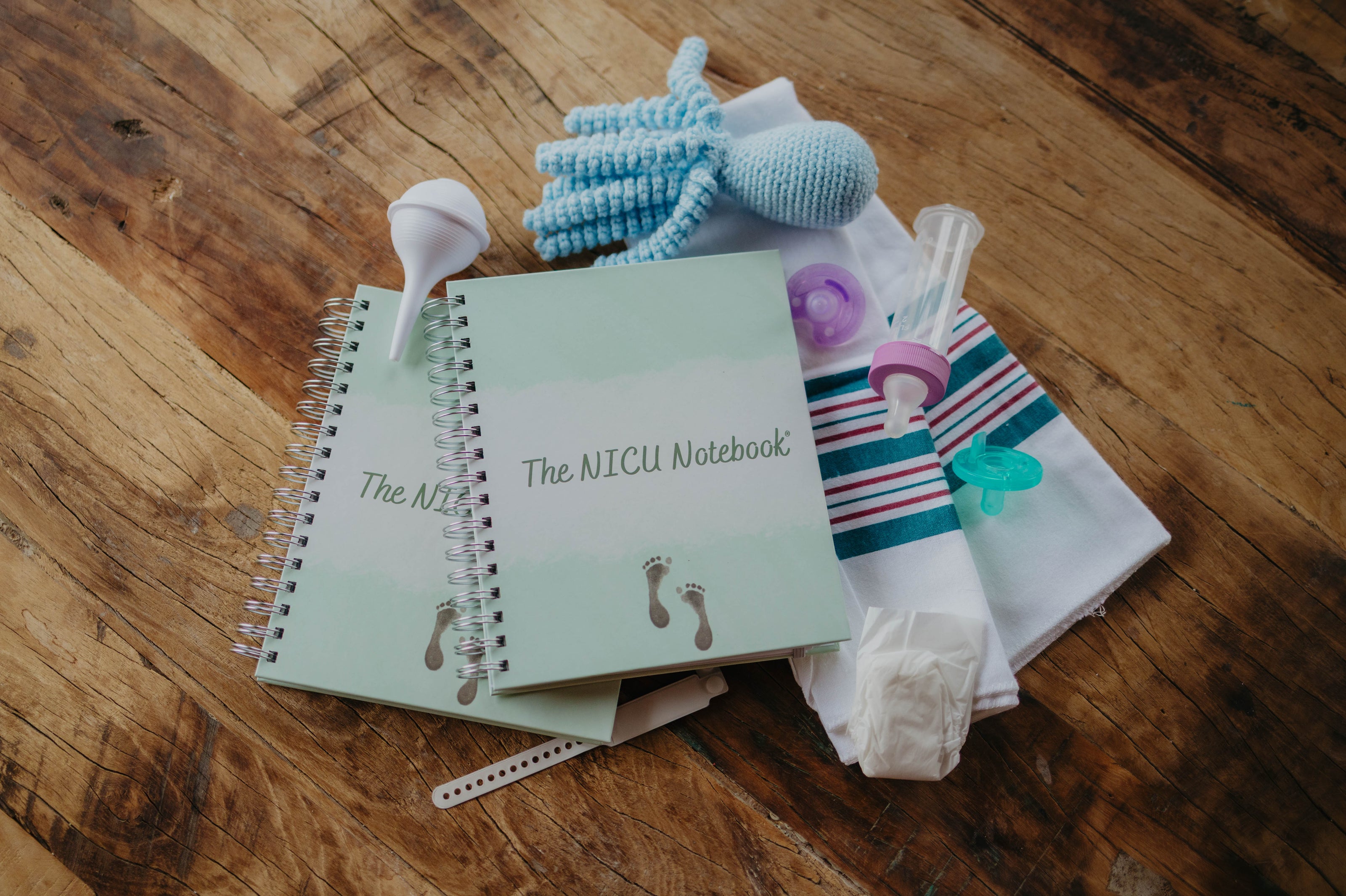 This image shows The NICU Notebook®, a coil bound NICU Journal for families with a baby in the Neonatal Intesive Care Unit, surrounded by baby items. Arranged on a wooden table top with the notebooks are a  blue crochet octopus, white nasal aspirator, purple binky, mint green pacifier, baby bottle, white micropreemie diaper, and blank neonatal hospital band.