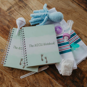 This image shows The NICU Notebook®, a coil bound NICU Journal for families with a baby in the Neonatal Intesive Care Unit, surrounded by baby items. Arranged on a wooden table top with the notebooks are a  blue crochet octopus, white nasal aspirator, purple binky, mint green pacifier, baby bottle, white micropreemie diaper, and blank neonatal hospital band.
