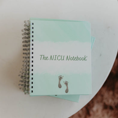  Photo taken from above of  The NICU Notebook®, a light green,coil-bound NICU Journal featuring a tiny micropreemie footprint on the cover placed on a white textured table.