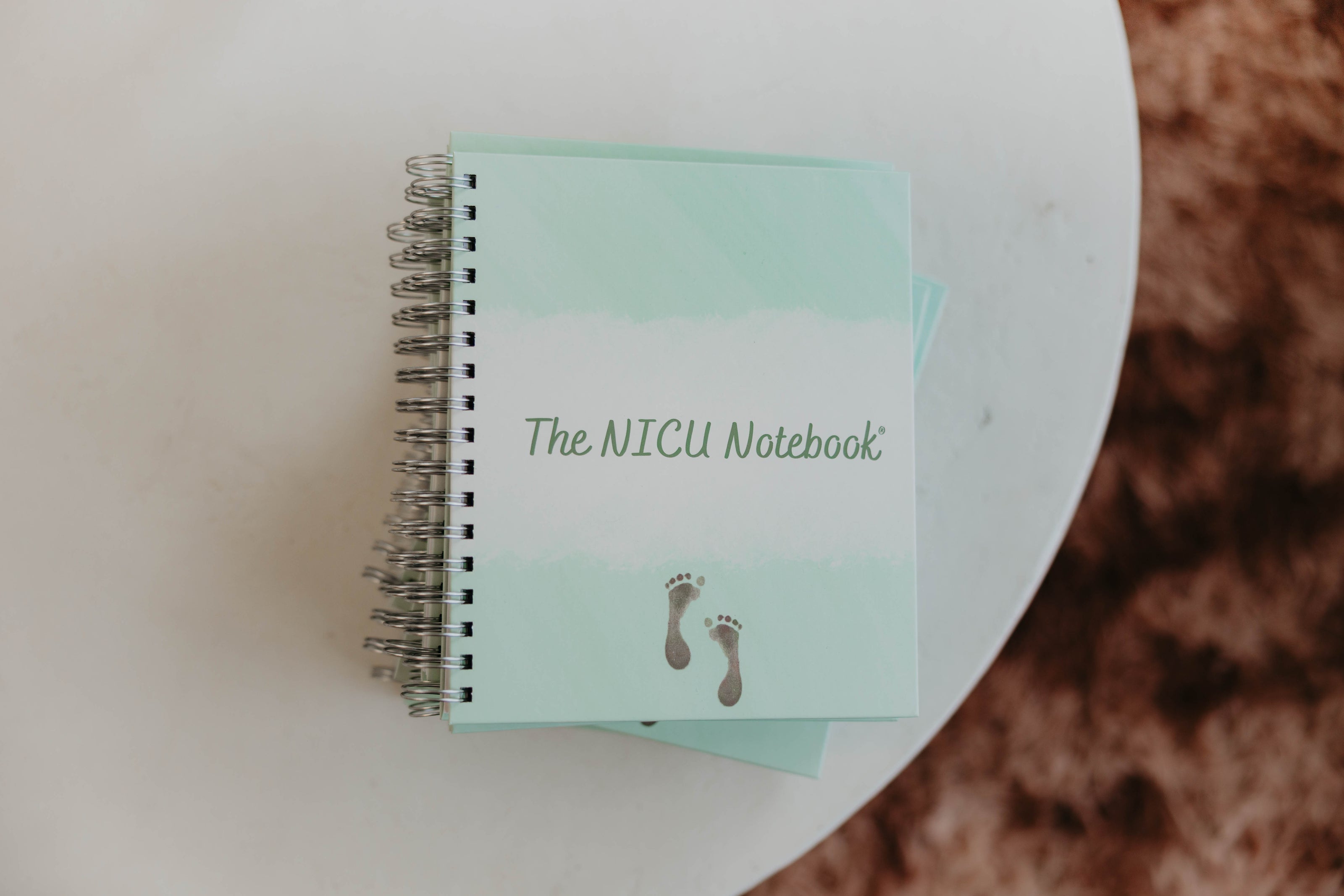  Photo taken from above of  The NICU Notebook®, a light green,coil-bound NICU Journal featuring a tiny micropreemie footprint on the cover placed on a white textured table.
