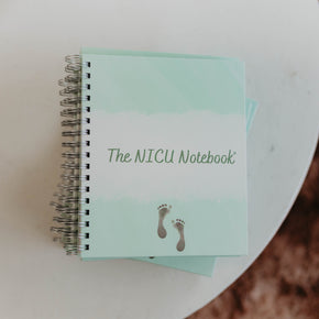  Photo taken from above of  The NICU Notebook®, a light green,coil-bound NICU Journal featuring a tiny micropreemie footprint on the cover placed on a white textured table.