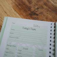 The NICU Notebook®, a light green, coil-bound NICU Journal, opened to a page from the Daily Journal section. The page features unfilled boxes that hold information such as 