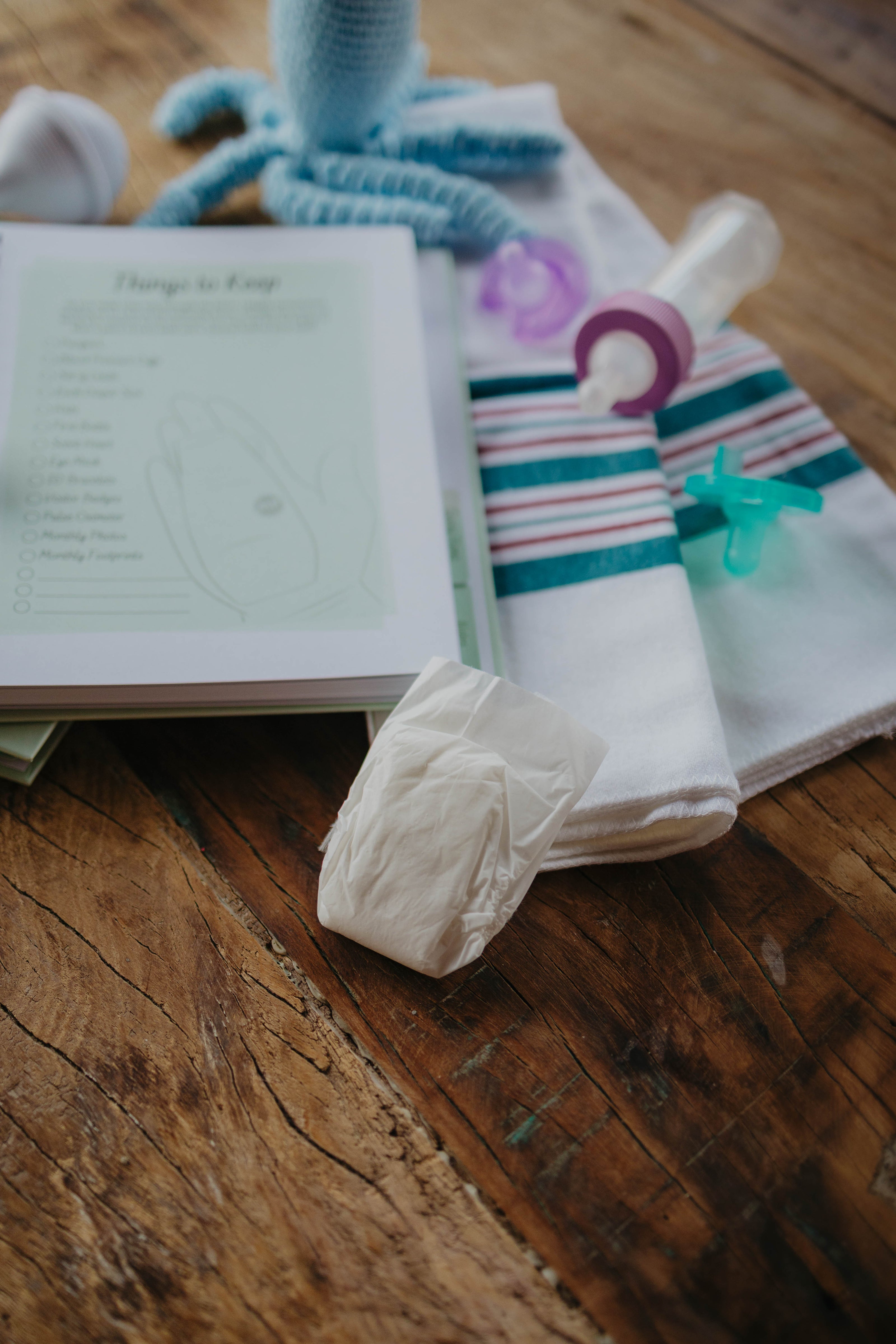 A  flat-lay of NICU baby essentials on a rustic wooden table, including a soft blue crochet octopus, a classic blue and pink newborn hospital blanket, a purple preemie pacifier and light green infant binky, a ready-to-feed single serve baby bottle, a white micropreemie diaper, and The NICU Notebook® opened to the 