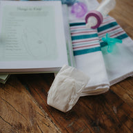 A  flat-lay of NICU baby essentials on a rustic wooden table, including a soft blue crochet octopus, a classic blue and pink newborn hospital blanket, a purple preemie pacifier and light green infant binky, a ready-to-feed single serve baby bottle, a white micropreemie diaper, and The NICU Notebook® opened to the 