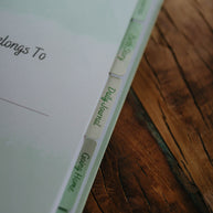 A close up of the section tabs in The NICU Notebook®, a light green, coil bound NICU Journal for families with a baby in the Neonatal Intesive Care Unit, on a wooden table top.